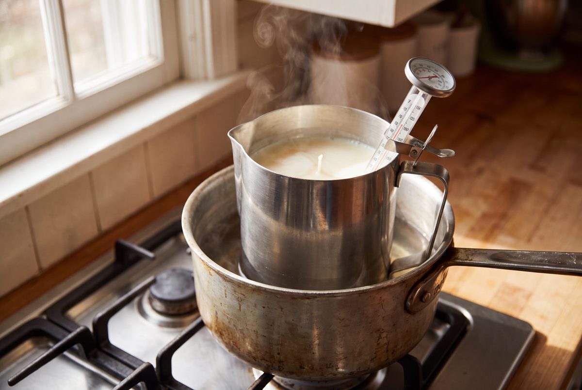 Soy wax melting in a metal pouring pitcher set in a saucepan of water, with a thermometer clipped to the pitcher.