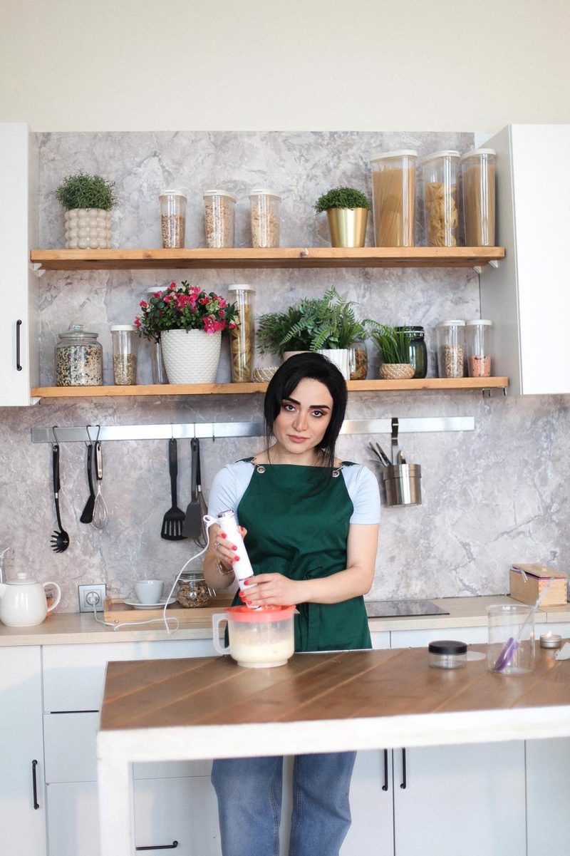 Nigar Rustamova at her workshop table, blending a batch with a hand blender, open shelves of jars and plants behind her.