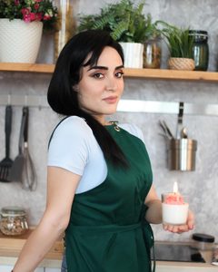Nigar Rustamova in a green apron in her kitchen workshop, holding a lit dessert candle in a glass.