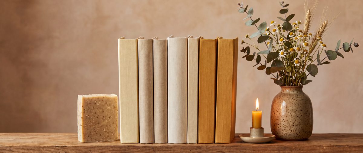 A row of linen-bound books on a wooden shelf with a handmade soap bar and a candle as bookends, and dried flowers in a vase.