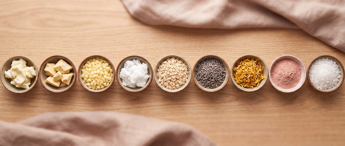 Overhead row of small bowls on light oak: shea butter, cocoa butter, beeswax pellets, coconut oil, oatmeal, lavender, calendula, pink clay and sea salt.