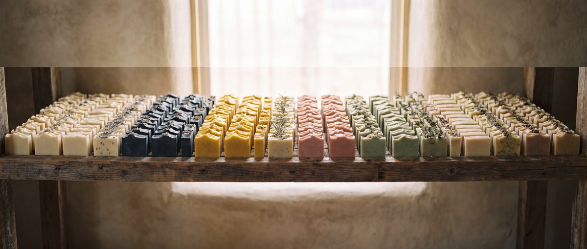 A long row of handmade soap bars curing on a wooden rack in cream, charcoal, turmeric, clay pink and sage, with herb sprigs between them.
