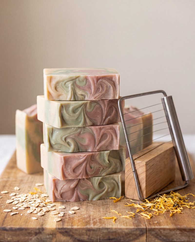 A stack of freshly cut soap bars with marbled swirls on a wooden board, with a soap cutter and scattered calendula petals.