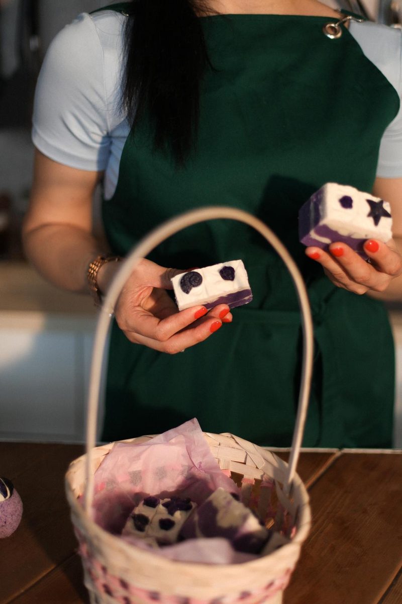 Nigar Rustamova holding two layered purple and white soap bars with embossed shells and stars over a basket of finished bars.