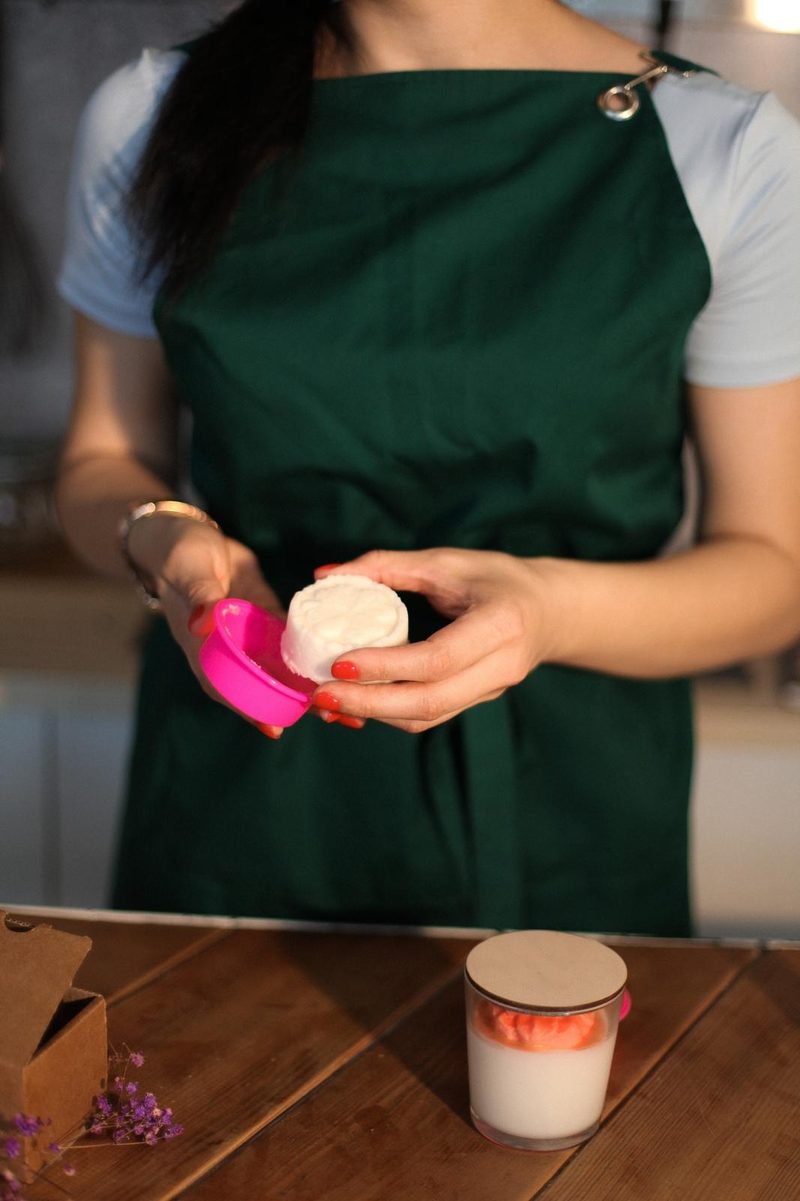 Hands releasing a white embossed bar from a pink silicone mould, a candle in a glass and a kraft box on the table.